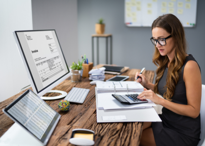 A brunette woman wearing glasses sitting at a wooden desk. She holds a pen in one hand while using the other to operate a calculator. The calculator rests on a ring binder folder.